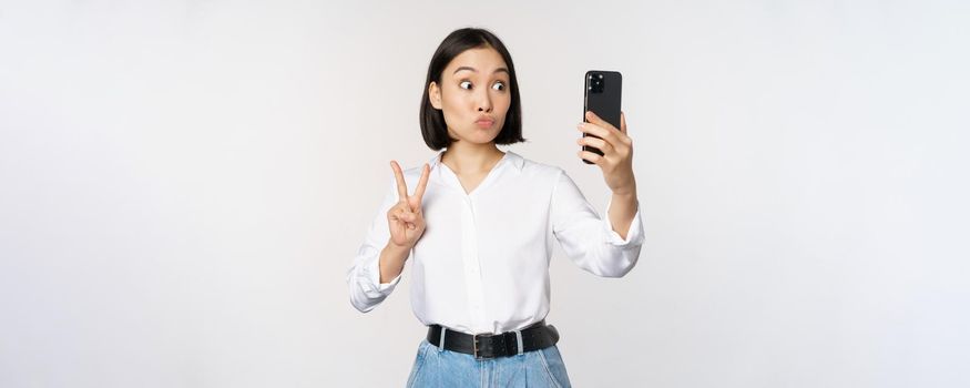 Funny asian girl showing peace, v-sign at smartphone camera, posing for photo, taking pictures with mobile application, standing over white background.