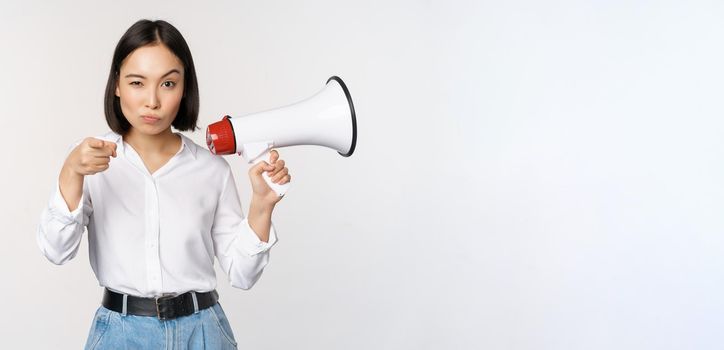 Image of modern asian woman with megaphone, pointing at you camera, making announcement, white background.