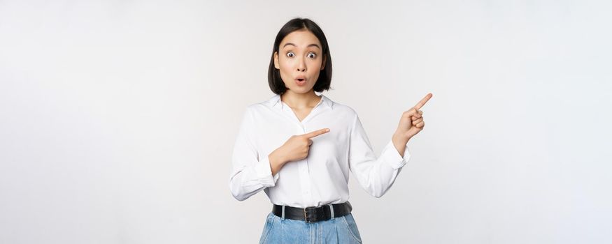 Surprised young woman pointing fingers right. Asian girl showing banner and looking enthusiastic, interested in advertising, standing over white background.