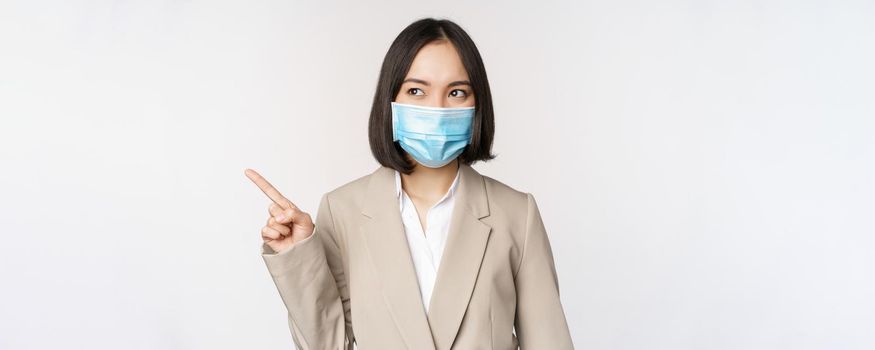 Enthusiastic businesswoman pointing fingers left, wearing medical face mask from covid-19 pandemic, standing over white background.