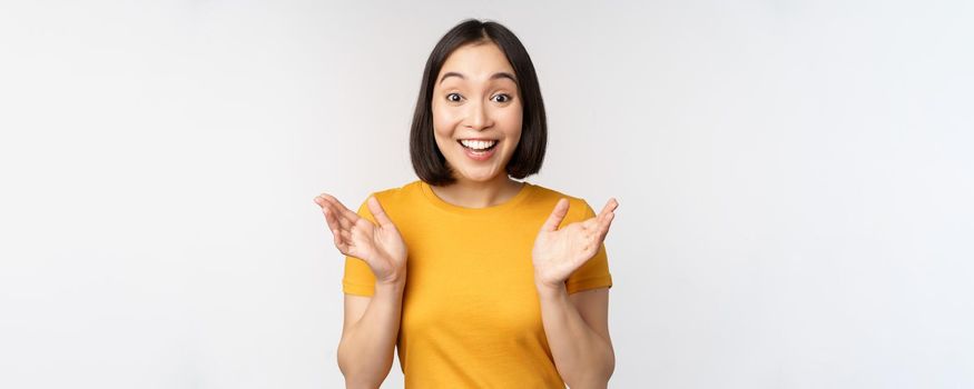 Close up portrait of asian woman looking surprised, wow face, staring impressed at camera, standing over white background in yellow t-shirt.