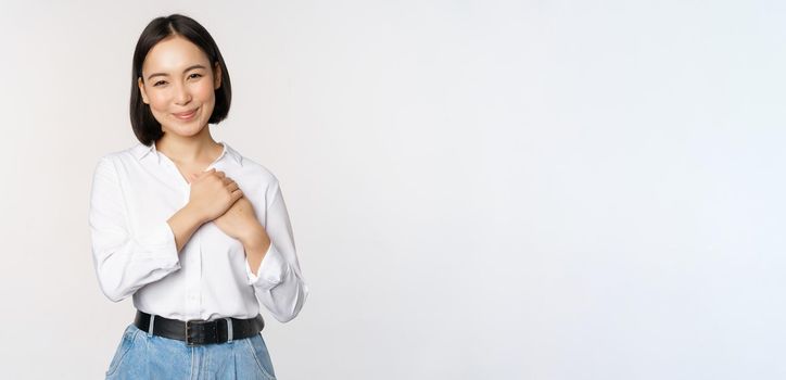 Image of caring young beautiful asian woman, holding hands on heart, looking pleased and happy, smiling at camera, standing over white background.