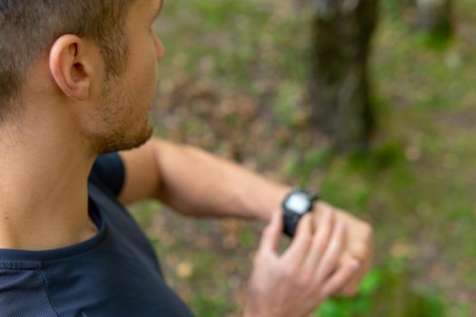 A man looks at the clock time in the park outdoors, around the forest, oaks green grass young enduring athletic athlete. active runner forest, jogger fit jog, woods outside. Autumn leisure feet stretches