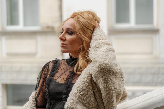 Profile of a beautiful blonde. She looks away and smiles. She is dressed in a black lace dress and a milky faux fur coat