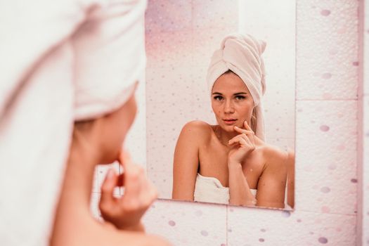 Portrait of young girl with towel on head in white bathroom looks and touches her face in the mirror and enjoys youth and hydration. Natural beauty, home care for problem skin.