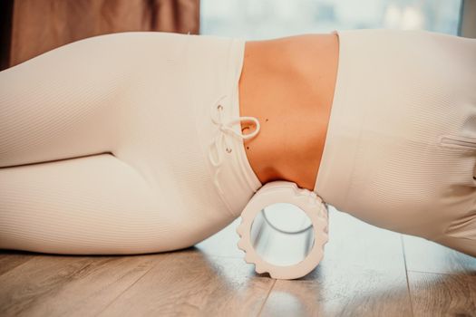 Adult athletic woman, in white bodysuit, performing fascia exercises on the floor - caucasian woman using a massage foam roller - a tool to relieve tension in the back and relieve muscle pain - the concept of physiotherapy and stretching training.