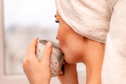 Young serene relaxed woman in spa bath towel drinking hot beverage tea coffee after taking shower bath at home. Beauty treatment, hydration concept