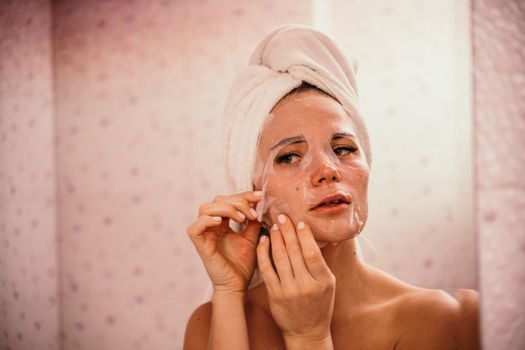 Young beautiful woman using a moisturizing facial mask after taking a bath. Pretty attractive girl in a towel on her head stands in front of a mirror in a home bathroom. Daily hygiene and skin care.