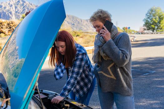 blonde adult woman and red-haired young woman looking at the engine of their broken down car with the bonnet open on the road while calling the emergency service, dressed in a blue jumper and a blue checked shirt.