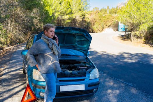 Modern and empowered blonde adult woman, dressed in blue jumper and blue jeans, standing on the road with the bonnet open repairing the engine of the car, while making a journey, on a sunny day with forest and a mountain in the background with cloudless blue sky.