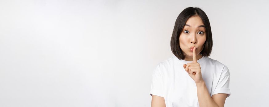 Close up portrait of young beautiful asian girl shushing, has secret, keep quiet silence gesture, press finger to lips, standing in tshirt over white background.