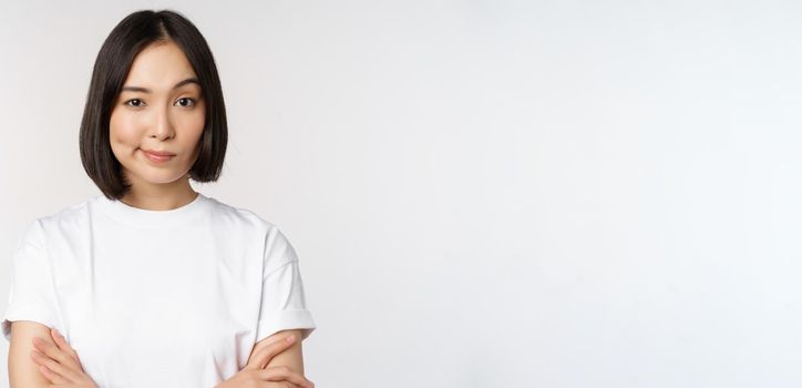 Close up of korean girl, looking skeptical, cross arms on chest and smirk, stare with disbelief at camera, standing over white background.