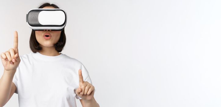 Potrait of asian woman in virtual reality glasses, pointing, choosing smth in VR headset, standing over white background.