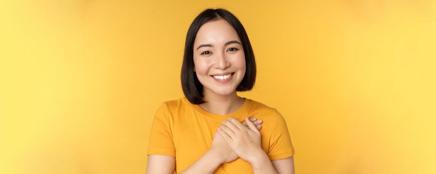 Beautiful asian woman, smiling with tenderness and care, holding hands on heart, standing in tshirt over yellow background.