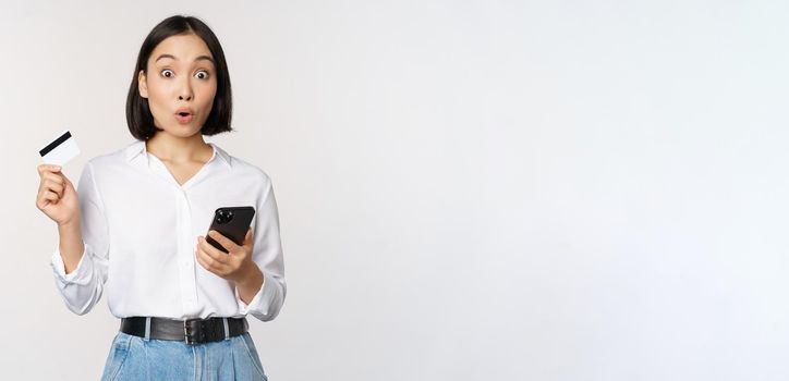 Online shopping concept. Image of surprised asian girl, holding credit card and smartphone, looking amazed in disbelief at camera, white background.