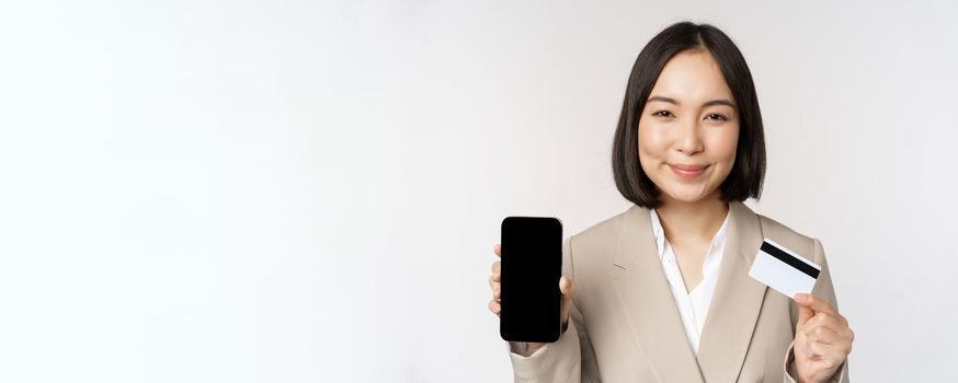 Smiling corporate woman in suit, showing mobile phone screen and app on mobile phone, smartphone screen, standing over white background.