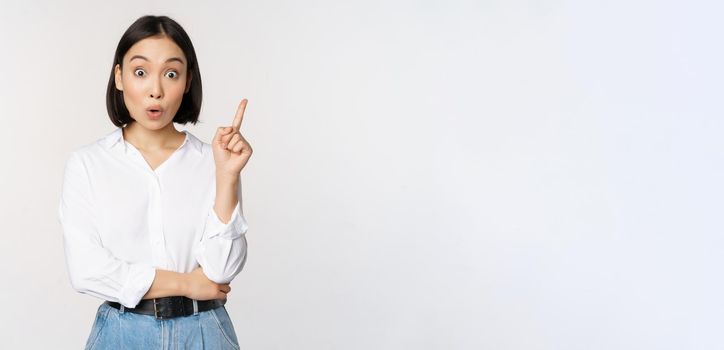 Eureka. Young smart asian girl has an idea, raising finger up, sharing her plan, pointing on top, standing over white background.