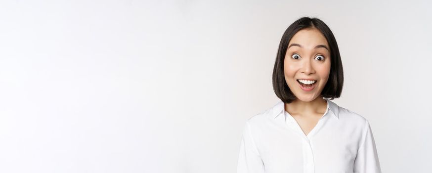 Image of korean woman looking surprised and happy at camera, standing over white background. Copy space