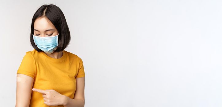 Covid-19, vaccination and healthcare concept. Cute asian girl in medical face mask, showing band aid after coronavirus vaccination, standing over white background.