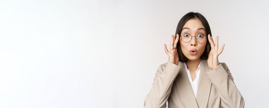 Portrait of asian businesswoman in glasses, looking surprised amazed at camera, standing in suit over white background.