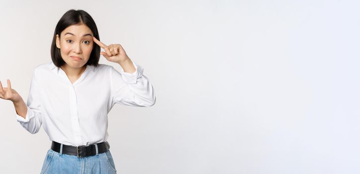 Are you insane. Portrait of frustrated young asian woman, roll finger near head and srugging, someone crazy or stupid, standing over white background.