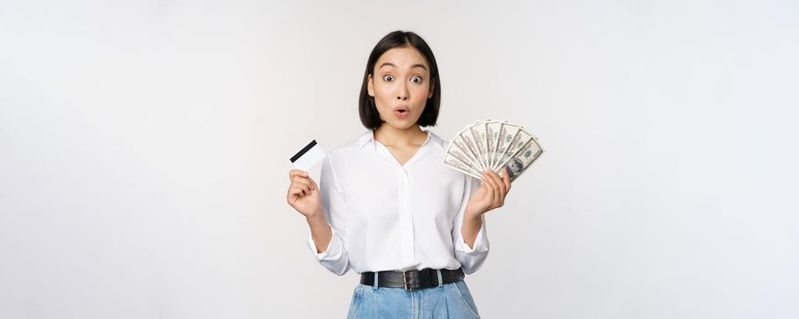 Finance and money concept. Happy young asian woman dancing with cash and credit card, smiling pleased, posing against white studio background.