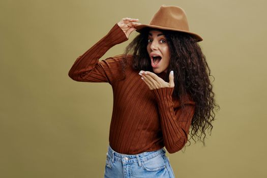 Wow idea. Enjoyed stylish curly Latin woman in brown hat, smiling at camera, holding hand on hat, closing mouth, isolated green background. Copy space clothing fashion brands, free place for your ad.