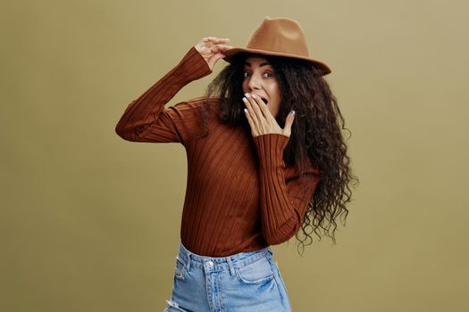 Wow. Enjoyed stylish curly Latin female in brown hat, smiling at camera, holding hand on hat, closing mouth, isolated green background. Copy space clothing fashion brands, free place for your ad.
