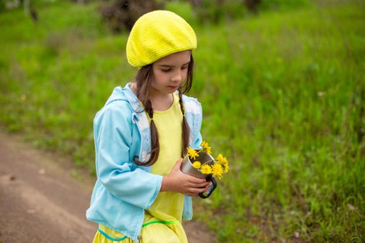 A little girl in a yellow knitted beret and a blue jacket walks along a path near a green lawn with a bouquet of dandelions in an iron mug