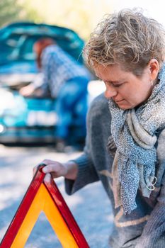 adult blonde woman in blue jumper in the foreground, putting a safety triangle on the road after an accident, on a sunny summer day with blue sky and natural light.