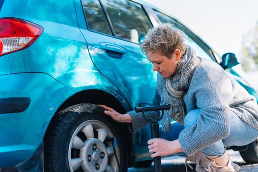 Adult blonde woman in blue jumper in the foreground, inflating the tyre of a blue car on the road after suffering a puncture while on a journey, on a sunny summer day with blue sky and natural light, with forest and mountain in the background.