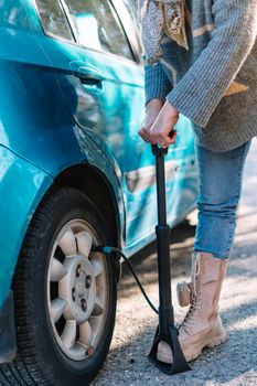 detail of a woman wearing a blue jumper, blue jeans and beige boots, inflating the tyre of her blue car after suffering a puncture on the back road, while on a journey.