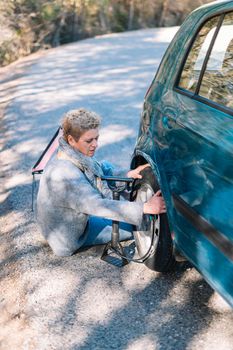 Adult blonde woman in blue jumper in foreground, inflating car tyre on the road after getting a puncture, on a sunny summer day with blue sky and natural light, with forest and mountain in the background.
