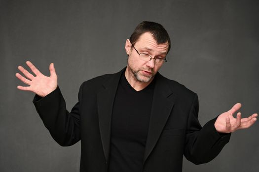 Businessman in a jacket and glasses with emotions talking to the camera, showing surprise. Portrait of an adult man on a gray background in the studio