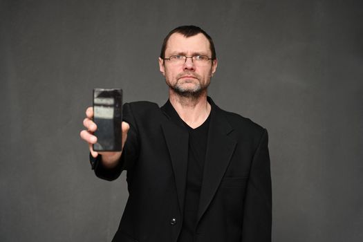 Caucasian businessman in a jacket looks at the camera and shows the display of a smartphone to the camera. Portrait of an adult man on a gray background in the studio