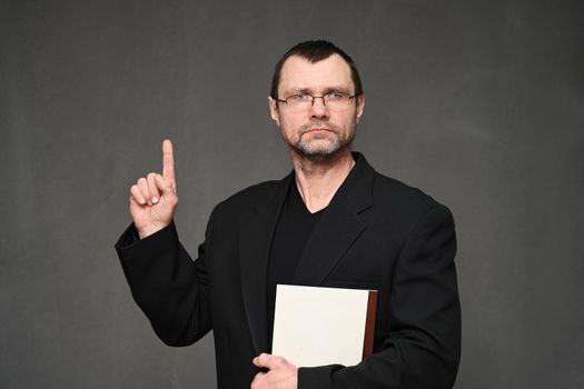 An adult man in a jacket looks at the camera with a folder in his hands, pointing his finger up. Portrait on a gray background in the studio