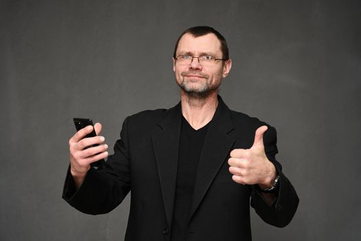 Caucasian businessman in a jacket looks at the camera with a smartphone in his hand and shows ok. Portrait on a gray background in the studio