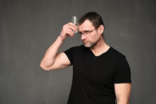 An adult man in glasses and a black T-shirt put a glass of water to his head on a gray background.