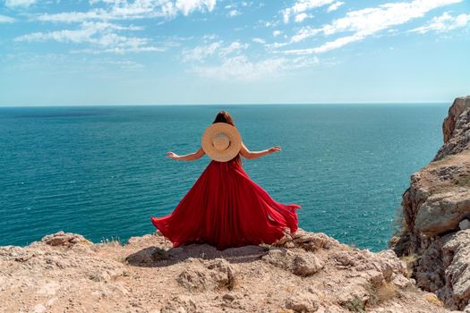 A woman, rear view in a red flying dress fluttering in the wind, a girl in a fluttering dress on the background of the sea. A straw hat hangs at the back of the neck