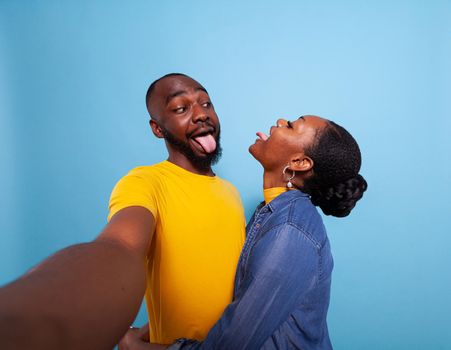 Silly lovers sticking tongue out at each other in front of camera, fooling around together in studio. Happy couple in love enjoying romance and affection, sharing relationship emotions.