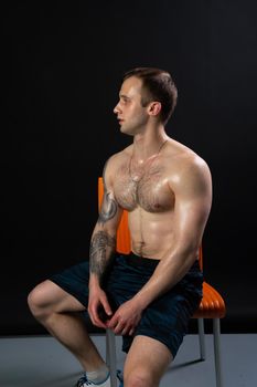 sits on an orange chair resting pumped Man on black background keeps dumbbells pumped up in fitness muscle torso, arm strong hold hand, healthy Attractive skin adult, one fit