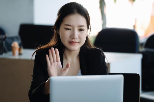 An Asian female employee or businessman is smiling and waving to a colleague using a notebook computer via video conferencing