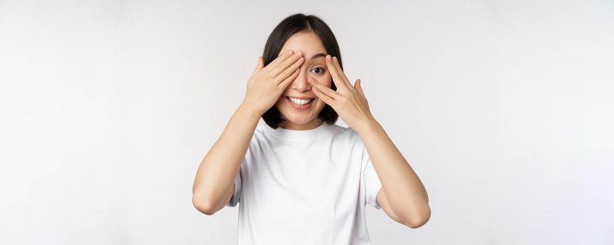 Portrait of asian woman covering eyes, waiting for surprise blindfolded, smiling and peeking at camera, standing over white background.