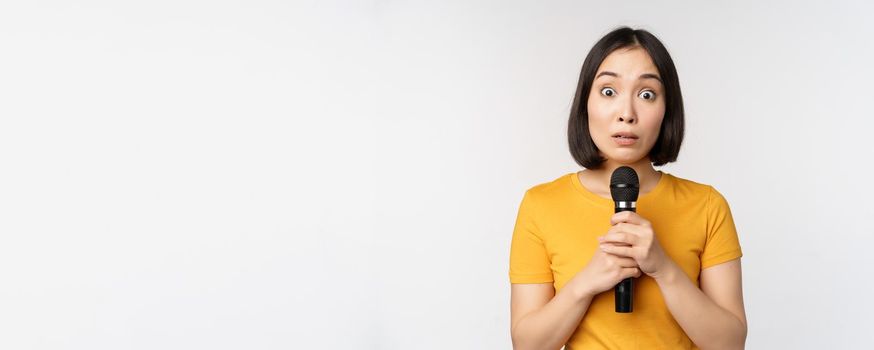 Modest asian girl holding microphone, scared talking in public, standing against white background. Copy space