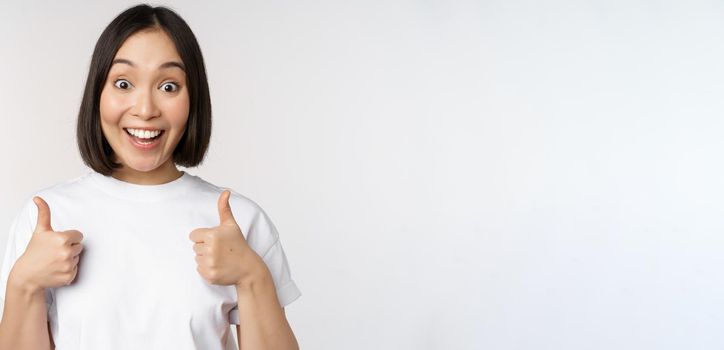 Happy young korean woman showing thumbs up and smiling, give positive feedback, recommending smth, standing over white background.