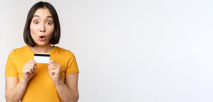 Portrait of beautiful korean girl holding credit card, recommending bank service, standing in yellow tshirt over white background.