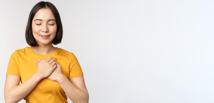 Romantic asian girfriend, holding hands on heart, smiling with care and tenderness, standing in yellow tshirt over white background.