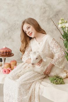 The girl is sitting at the Easter table with cakes, spring flowers and quail eggs and holding a white kid in her arms. Happy Easter celebration