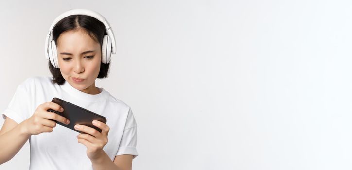 Happy asian woman in headphones, looking at smartphone, watching video on mobile phone and smiling, standing over white background.