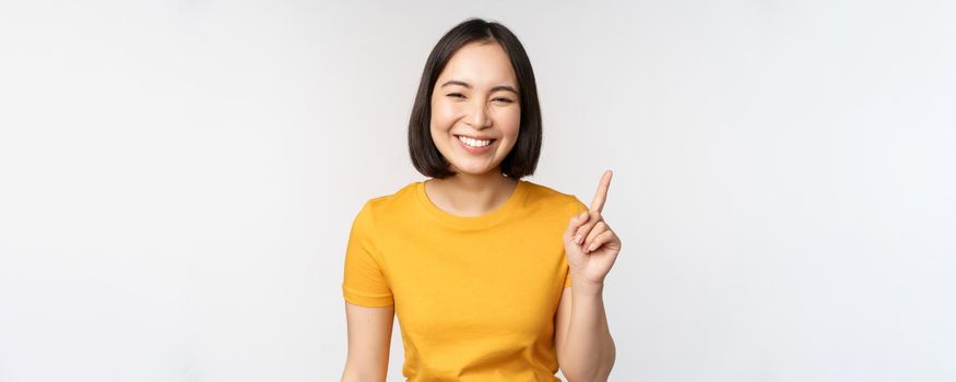 Beautiful young asian woman pointing finger up, smiling and looking amused at camera, showing advertisement, announcement on top, white background.